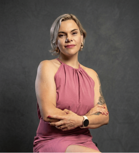 #ParaCegoVer: Dan&uacute;bia Don&aacute;, mulher de cabelos loiros curtos, sorrindo com seguran&ccedil;a. Usa vestido rosa com decote halter e smartwatch. Bra&ccedil;os cruzados, postura confiante. Fundo cinza escuro. Foto profissional de Consultora de Beleza Independente Mary Kay.